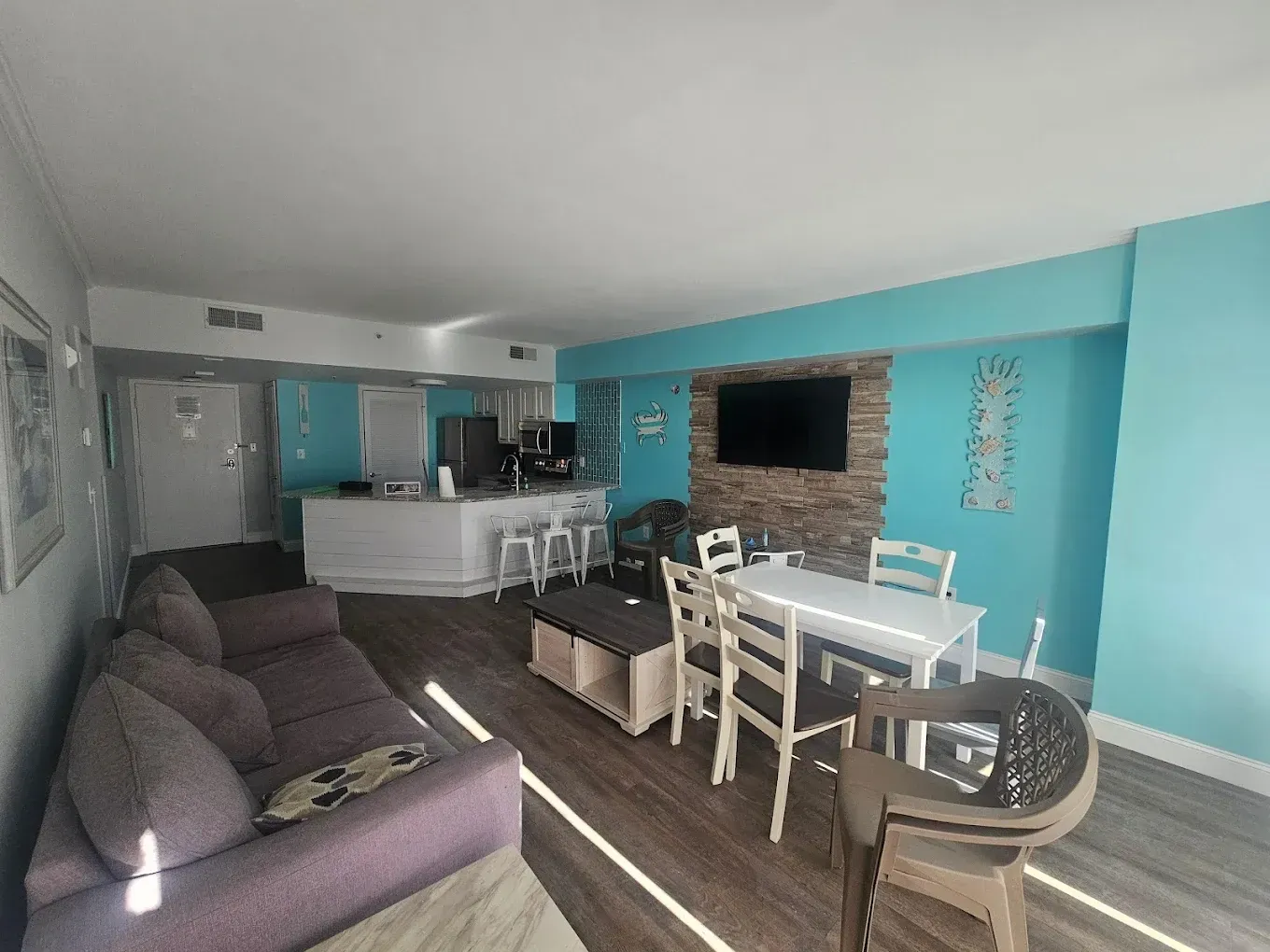 Living area with a purple couch, dining table, and kitchen in a condo. Turquoise walls, wood flooring.