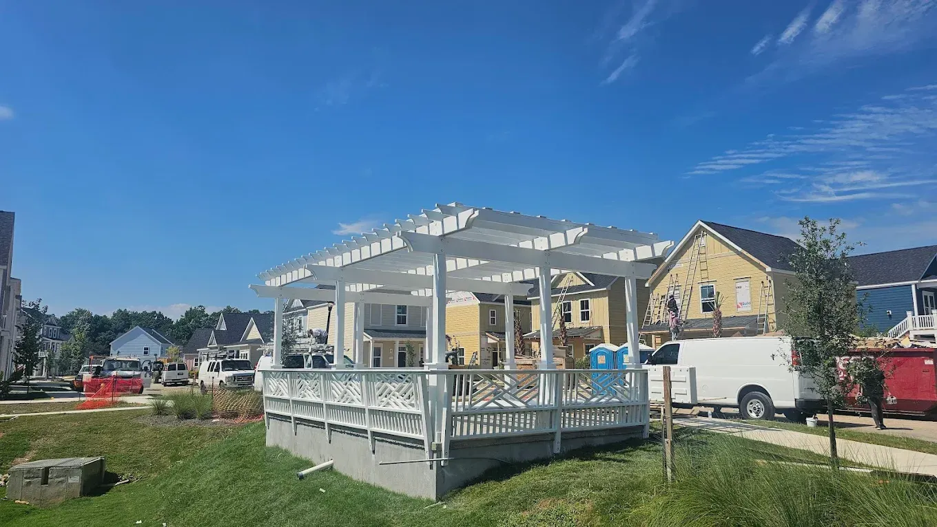 White pergola on a concrete base, over a wooden deck, with houses and blue sky in the background.