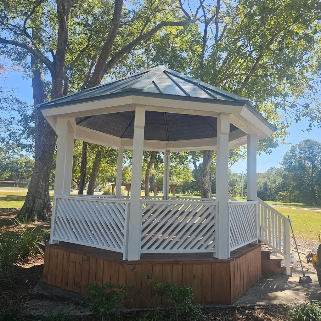 White gazebo with green roof, angled lattice railing, and wooden base in a park setting.