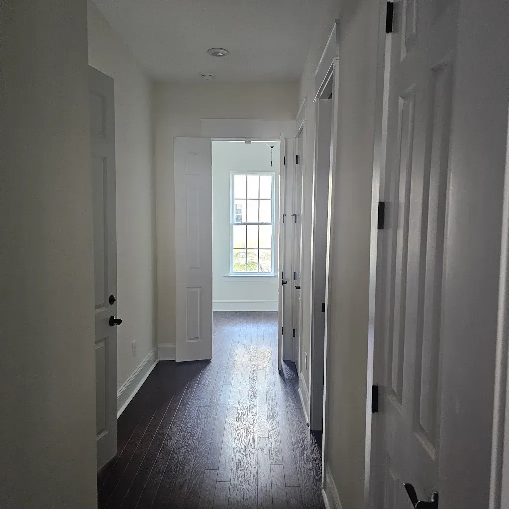 Hallway with dark wood floor, white walls and doors, and a window at the end.