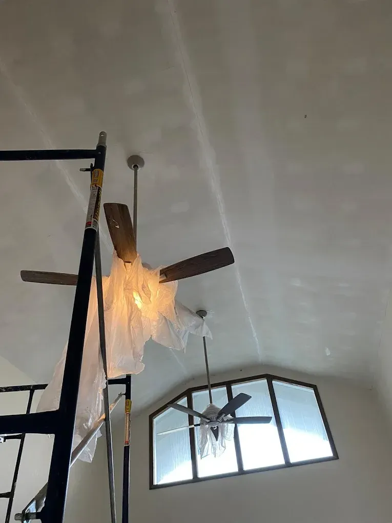 Scaffolding next to ceiling fan covered in plastic. Another fan and arched window visible. White ceiling.
