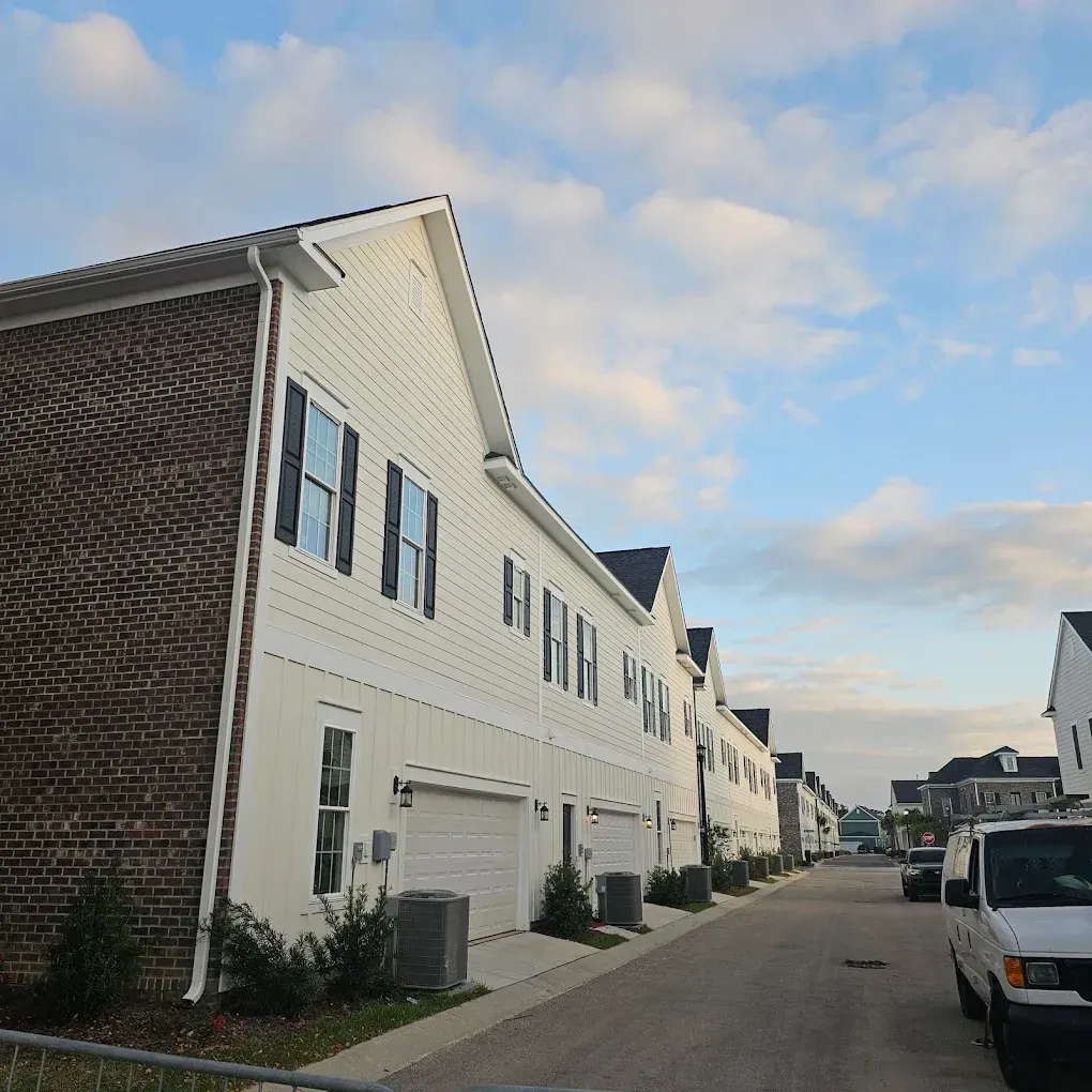 Row of white townhouses with black shutters, brick accents, and attached garages on a street. Blue sky.