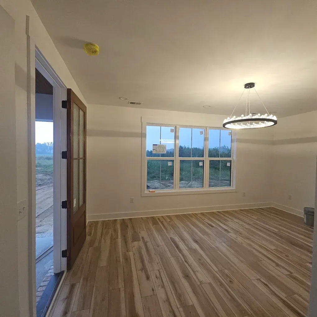 An empty room with wood floors, large windows, and a chandelier. A door is on the left.