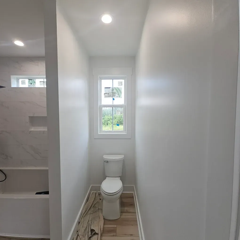 Narrow bathroom with white walls, toilet, small window, and wood-look tile floor.