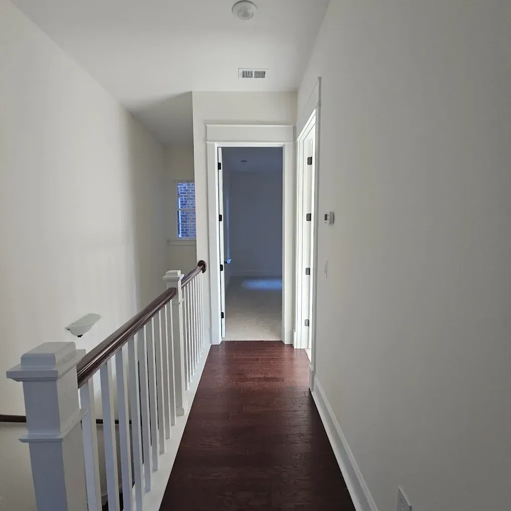 Hallway with wood floor and white walls, leading to open doorway and stairs with railing.
