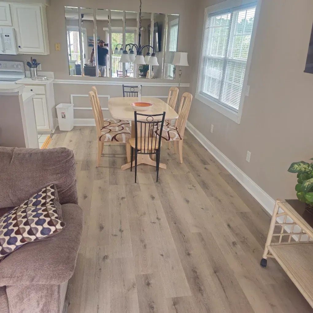 Dining area with wood flooring, table, chairs, and window. Beige walls. Brown sofa.