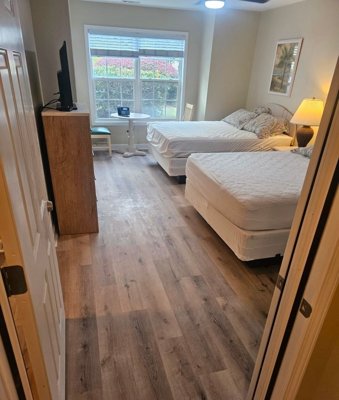 Narrow hallway with dark wood floors, white walls, and closed doors. A window is at the end of the hall.