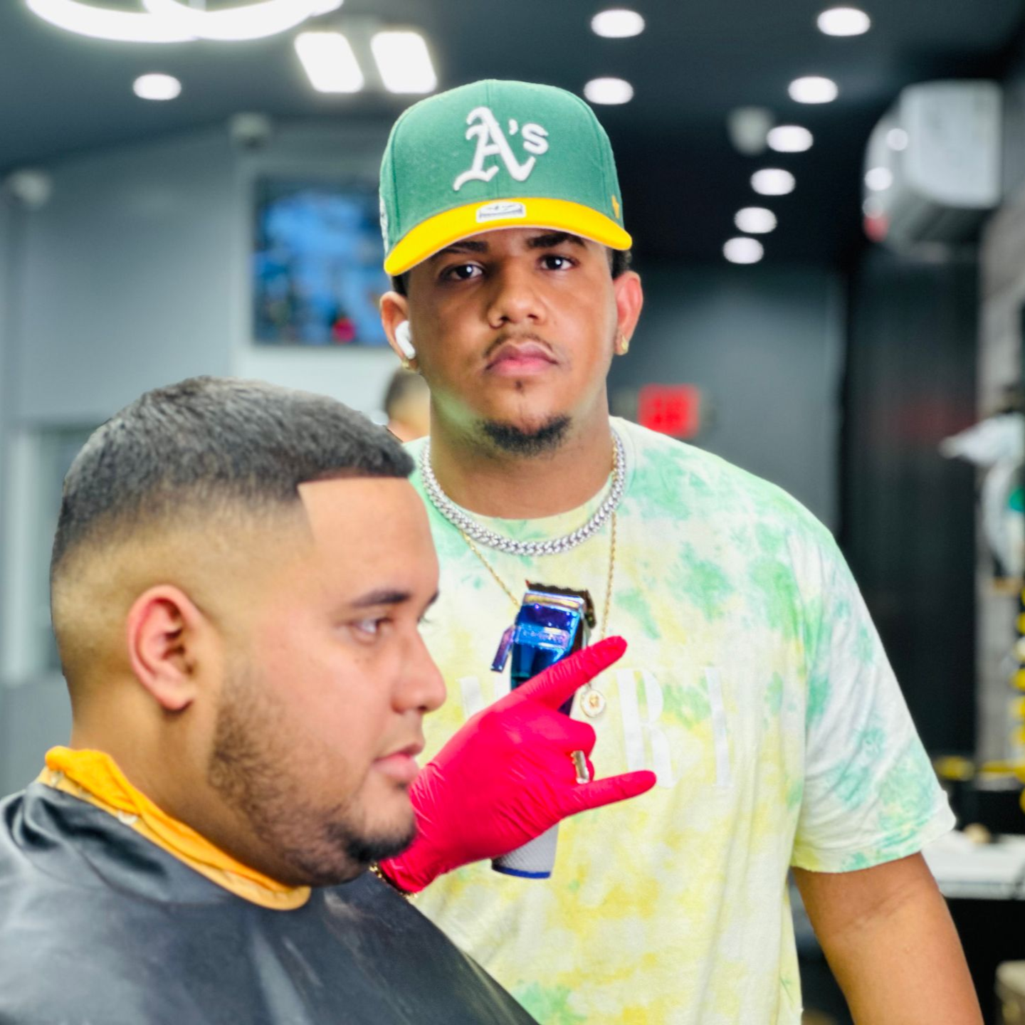 Two men in a barbershop, one in a green A’s cap, the other in a red glove holding a phone.