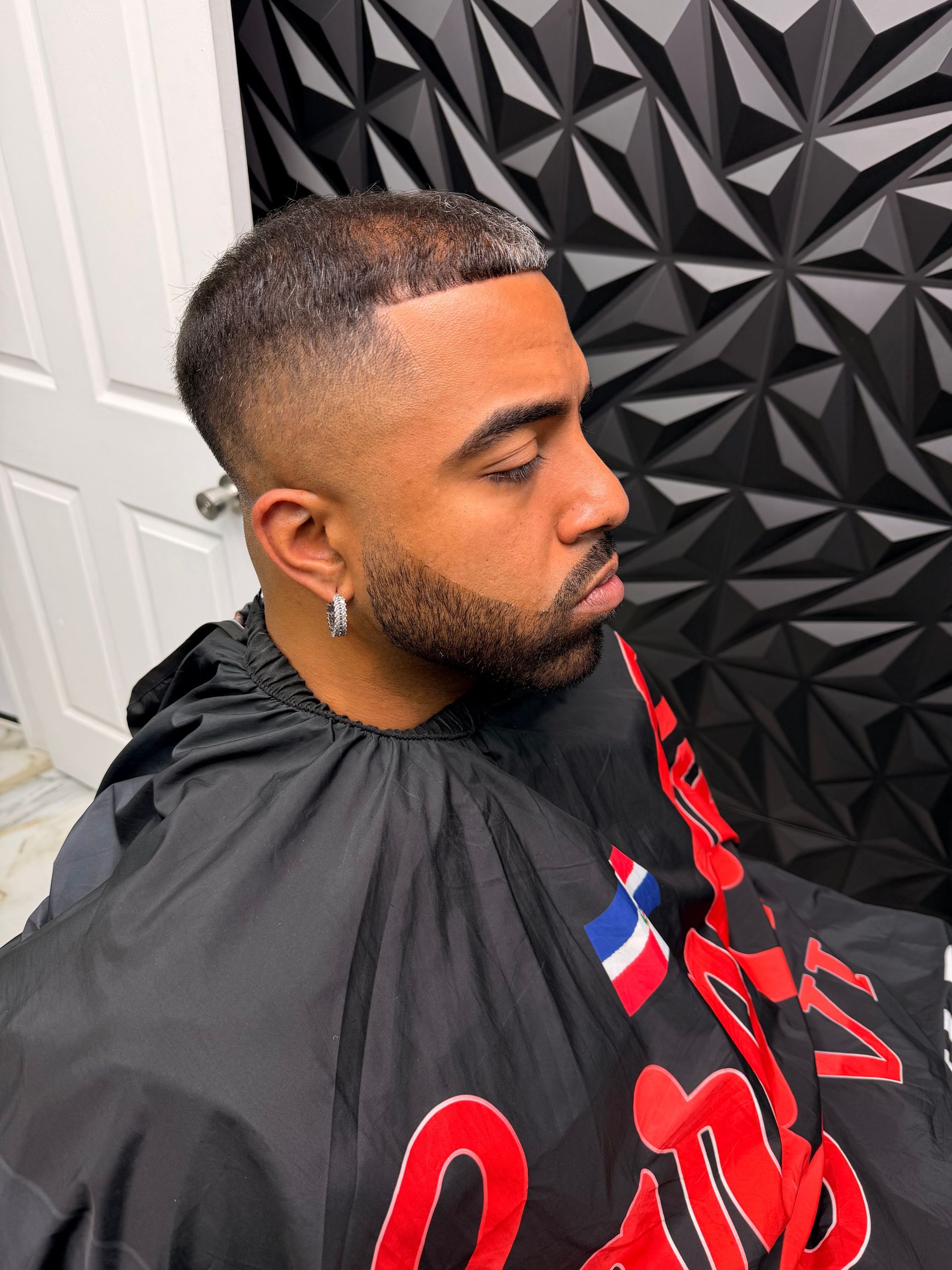 Close-up of a man with a fade haircut and beard, wearing a black barber cape in a barbershop
