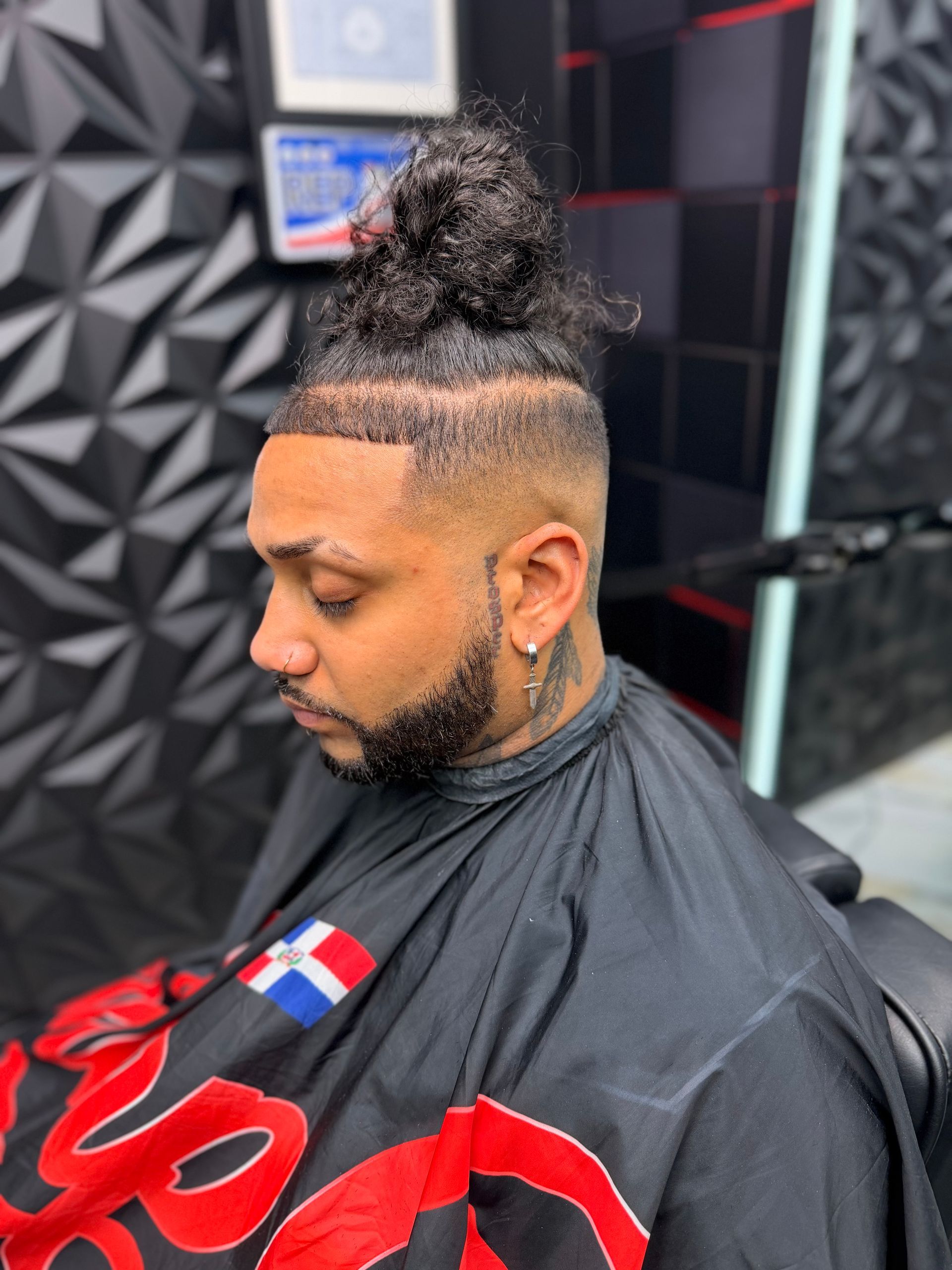 Man with curly topknot haircut and beard seated in barber chair, wearing black cape in a barbershop