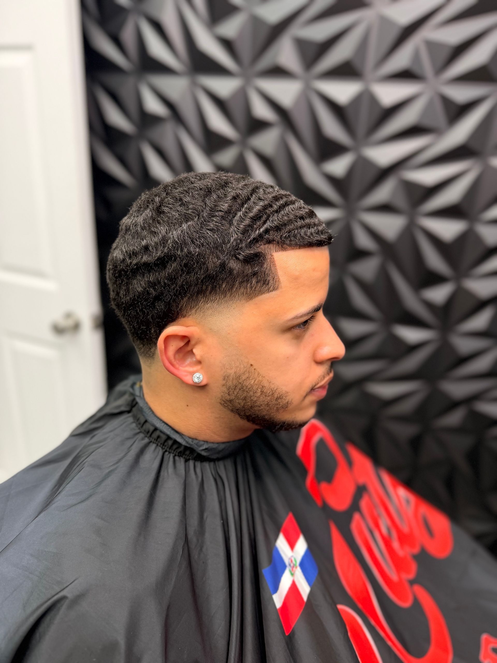 Man getting a haircut in a barbershop, side profile with a patterned black cape and geometric wall decor.