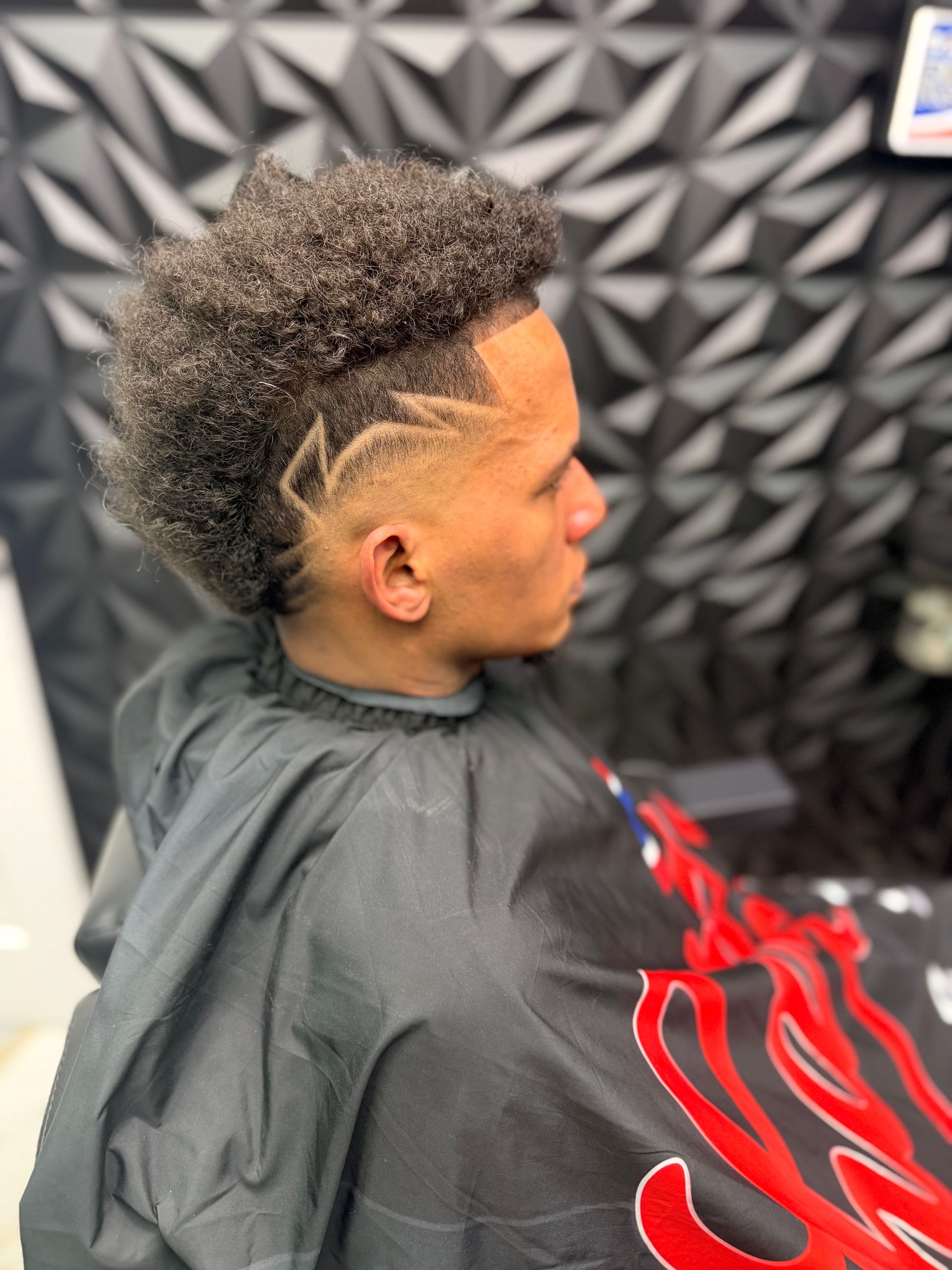 Side-profile haircut with a curly top fade and shaved design, draped in a black barber cape.