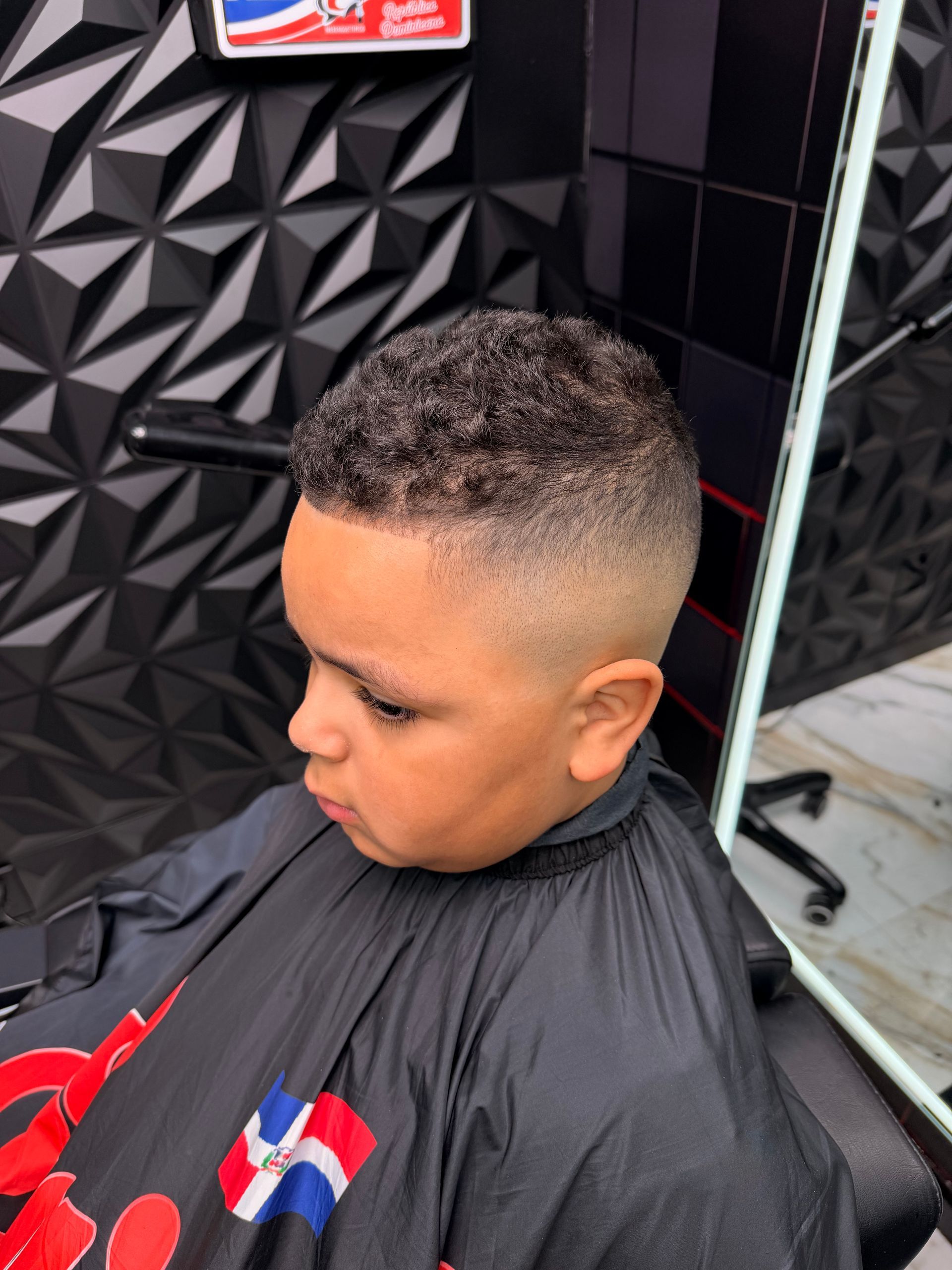 Boy with a short fade haircut sitting in a barber chair, wearing a black cape in a barbershop.