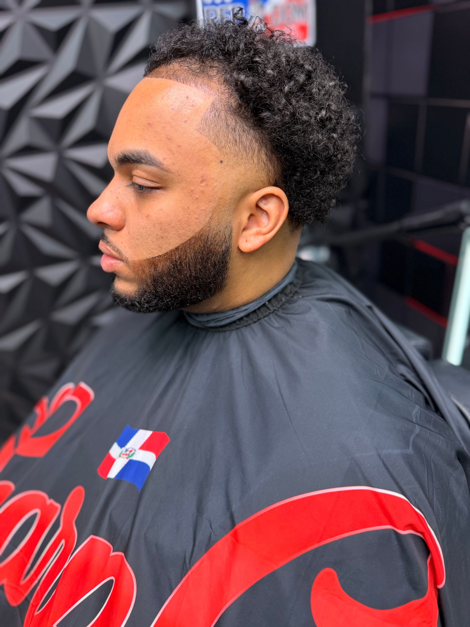 Profile view of a man in a black barber cape with red lettering, seated in a barbershop.