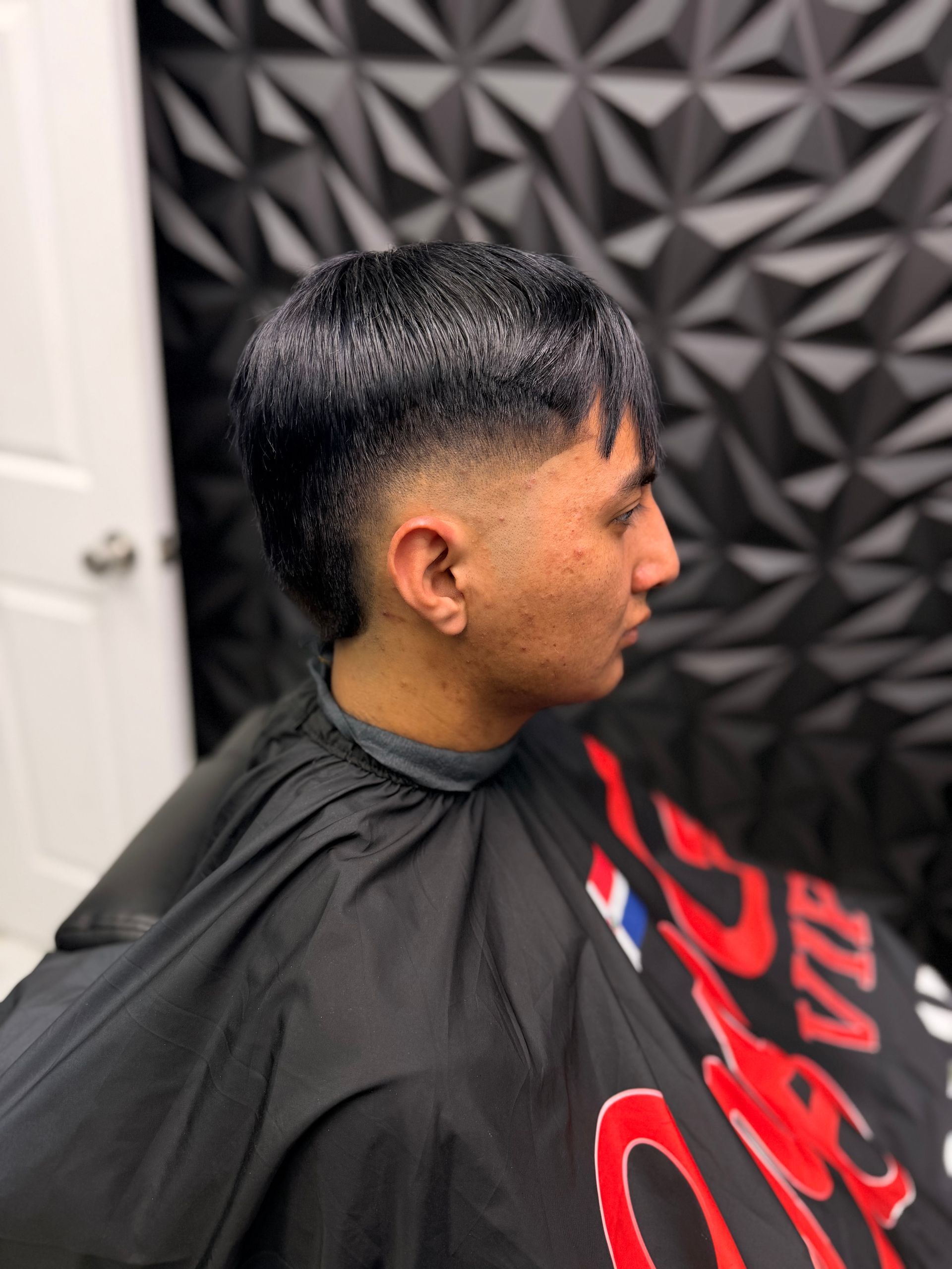 Side-profile haircut in a barber chair, with a black cape and a textured gray wall background