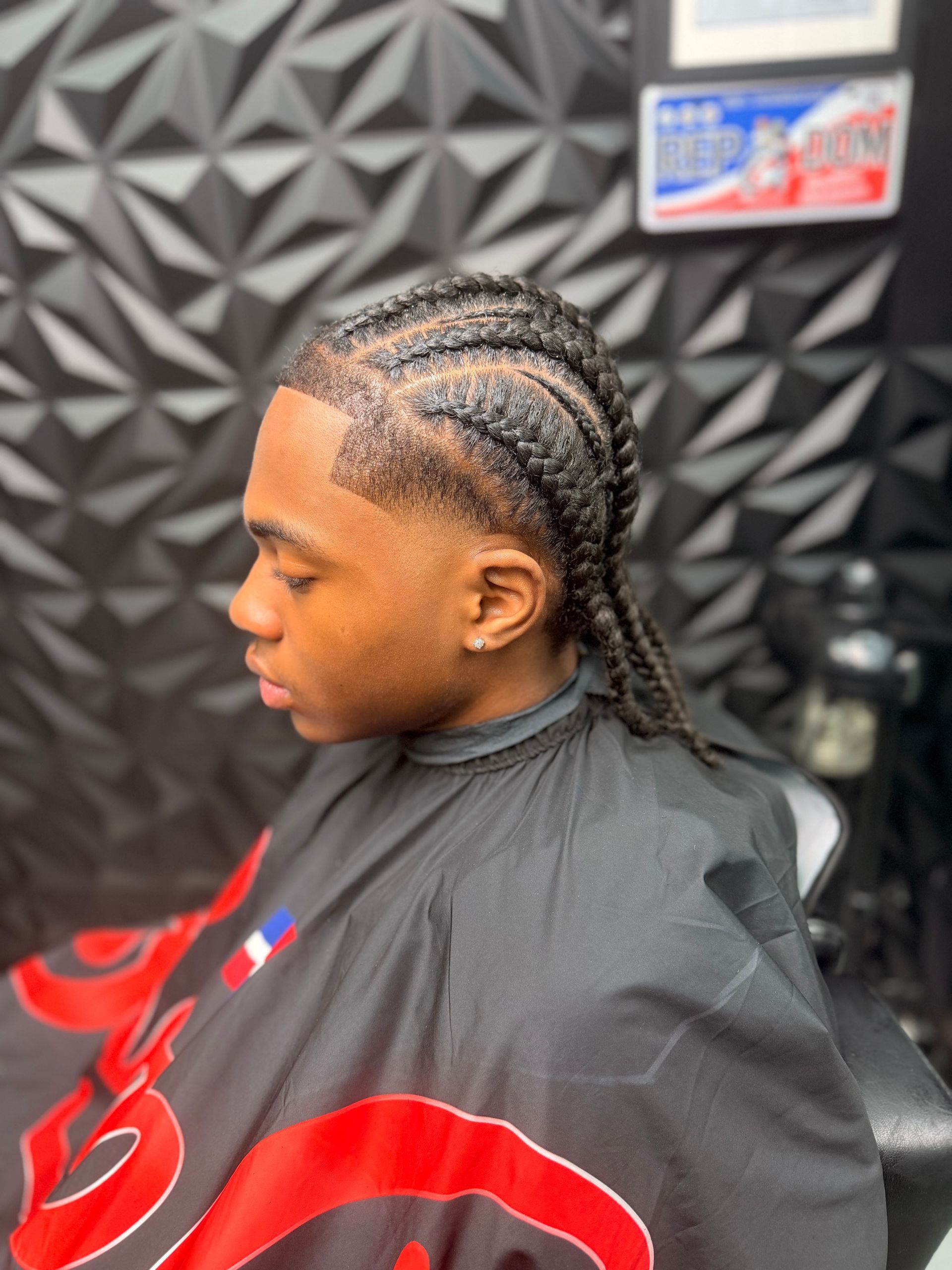 Person with braided hair seated in a barber chair, wearing a black cape with red designs, in a barbershop