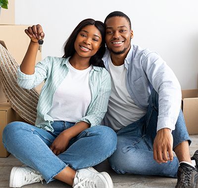 Couple seated, smiling, holding house keys near moving boxes.