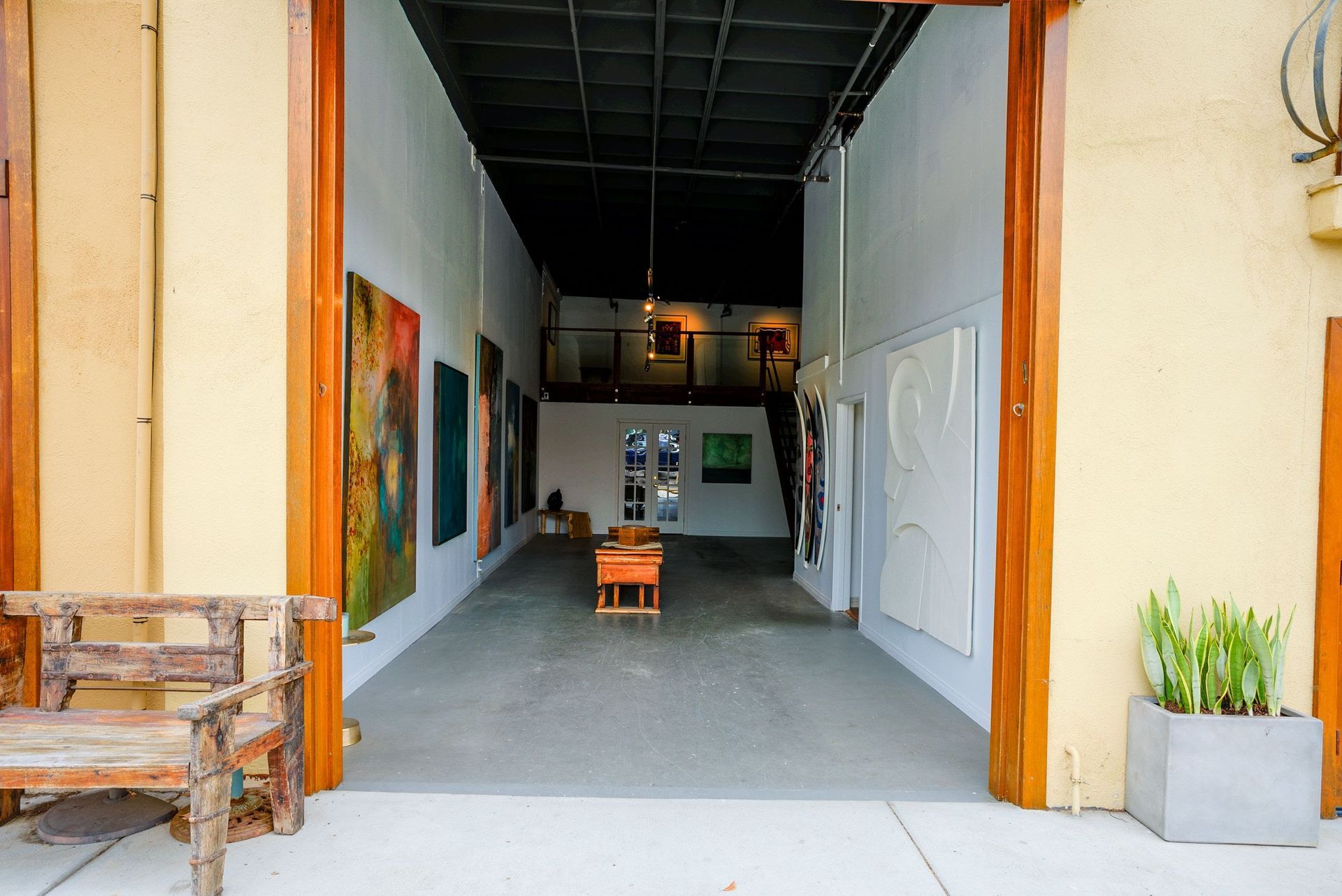 The entrance to Palm Palm Art Collective. Looking into a large room with a lot of paintings on the walls and a bench in the middle of it .