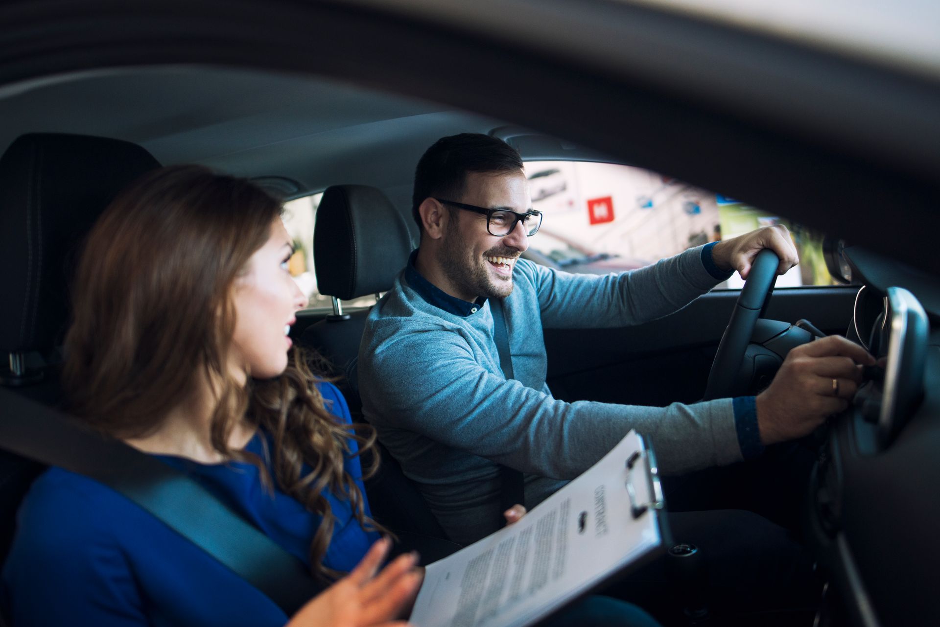 Dos personas en un coche, sonriendo y charlando mientras una conduce y la otra sostiene un documento.