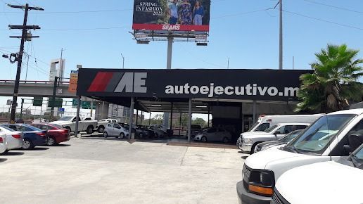 Estacionamiento de concesionario de autos con filas de autos y un letrero negro “autoejecutivo.mx” bajo un cielo despejado.