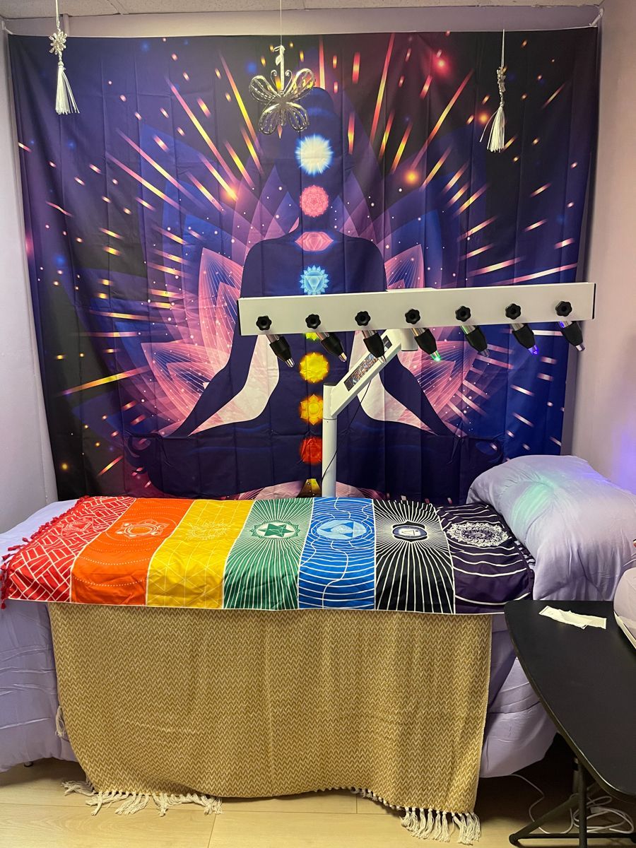 A wellness room with a chakra-themed tapestry, light therapy machine, and rainbow towels on a massage table.