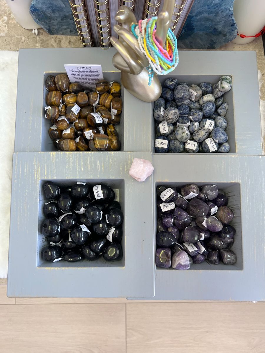 Four squares of gemstones: tiger's eye, gray, black, purple. A pink crystal and silver hand sculpture are also present.