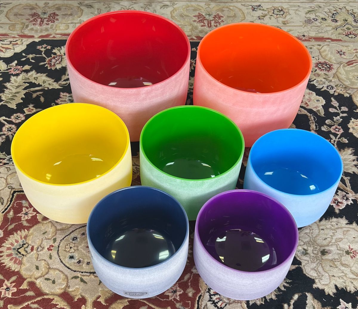 Seven colored crystal singing bowls on a patterned rug; each bowl in a different color of the rainbow.