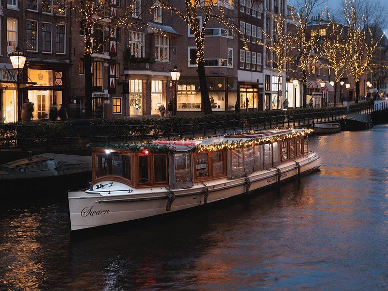Boat on Amsterdam canal at dusk, lit with Christmas  marketlights; buildings line the canal.