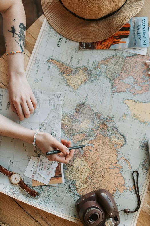 Hands pointing at a world map, planning travel. Items include a hat, camera, and watch.