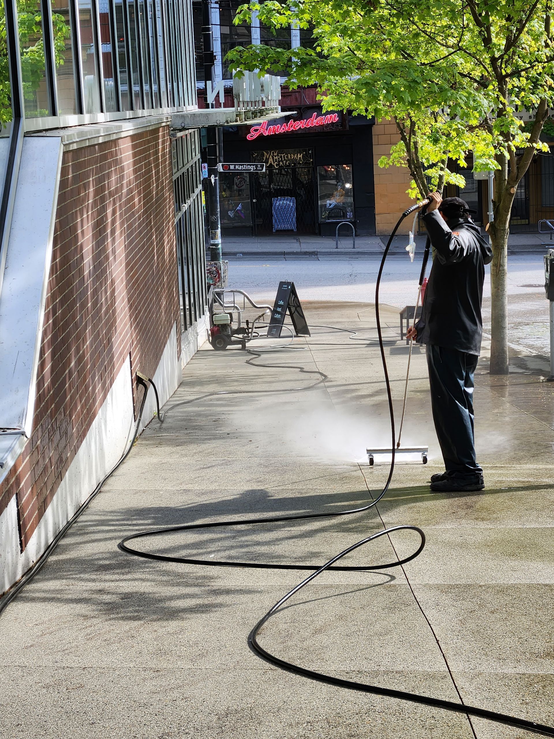 Person pressure washing a sidewalk in front of a brick building with graffiti and a business in the background.