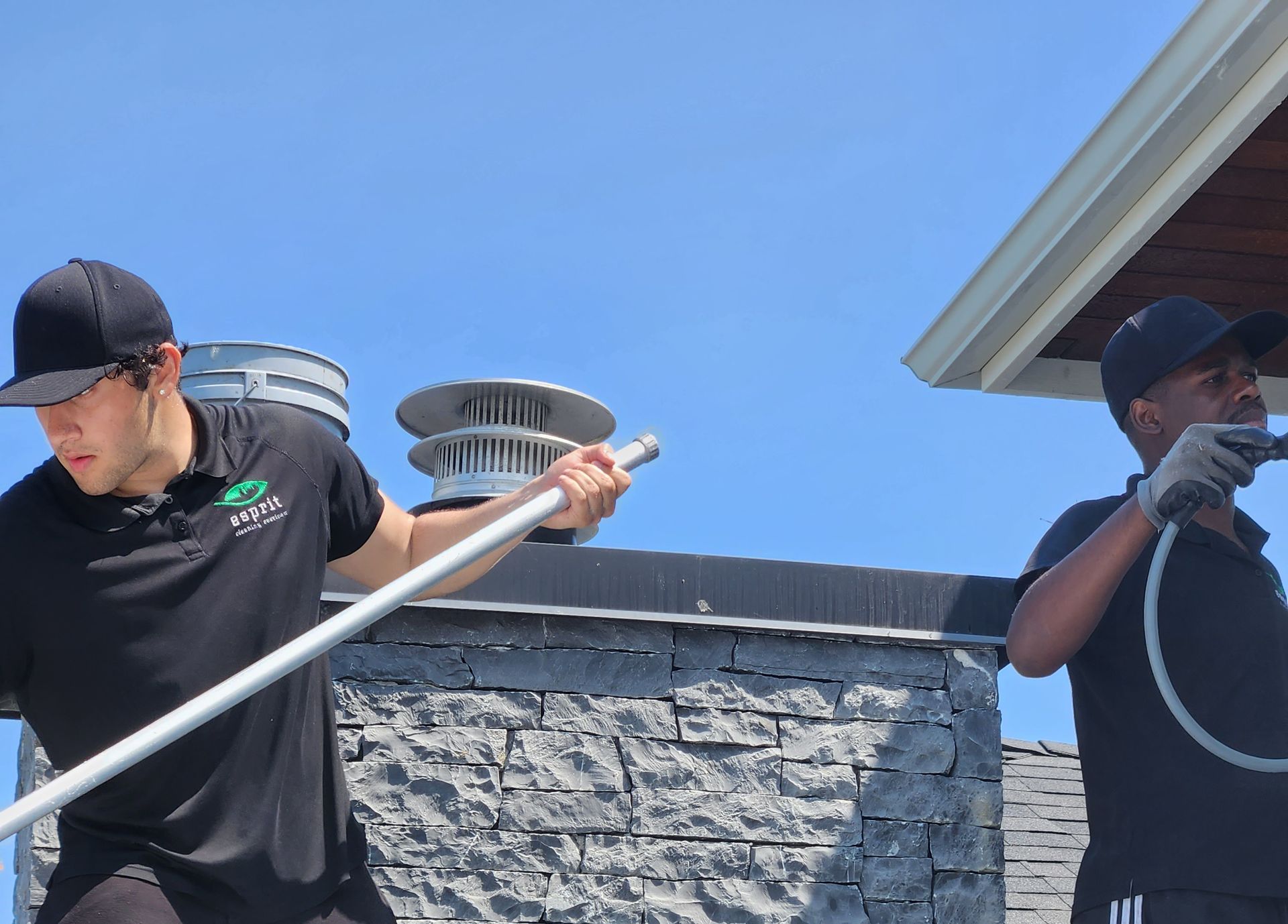 Two workers in black uniforms perform maintenance on a stone chimney exterior under a clear blue sky.