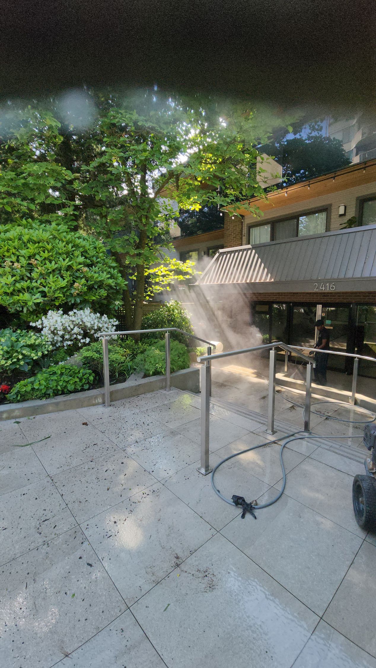 Misting system on a patio near lush greenery and a building with dark windows. Sunlight and hazy.