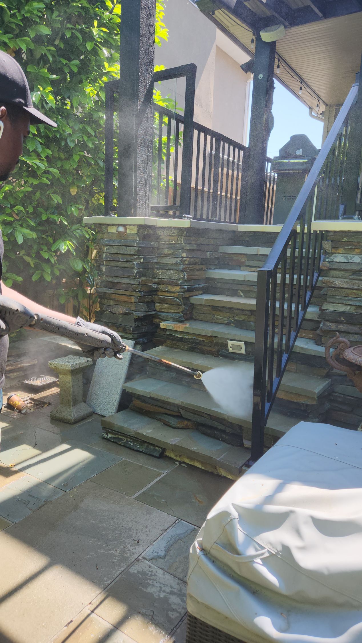 A person pressure washes stone steps and a wall on a sunny patio.