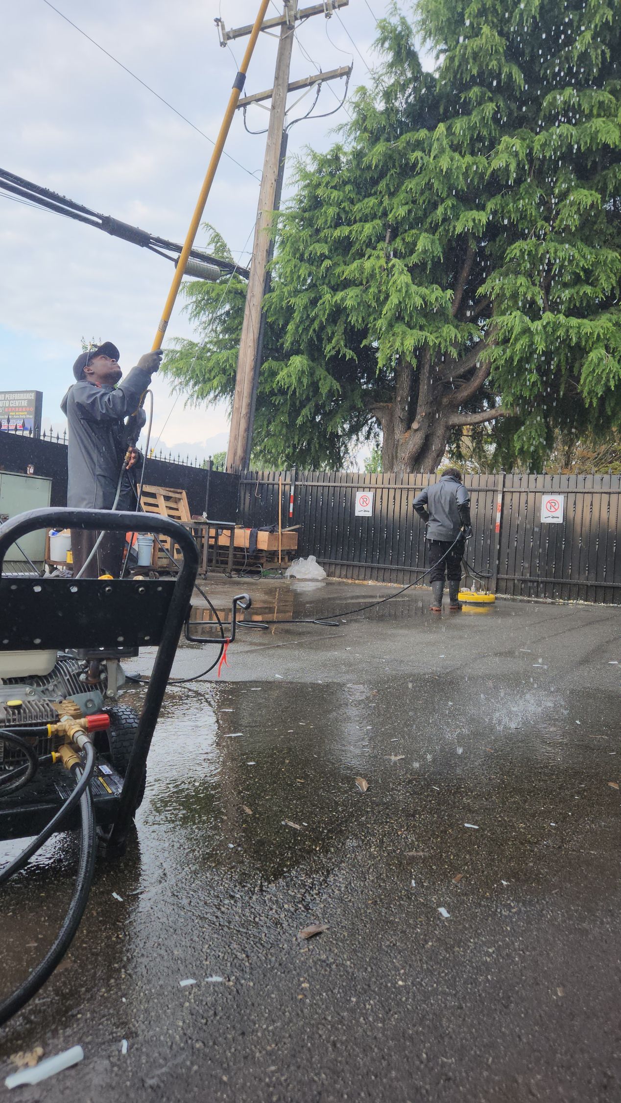 Two workers using pressure washers to clean a dirty road with utility poles in the background.