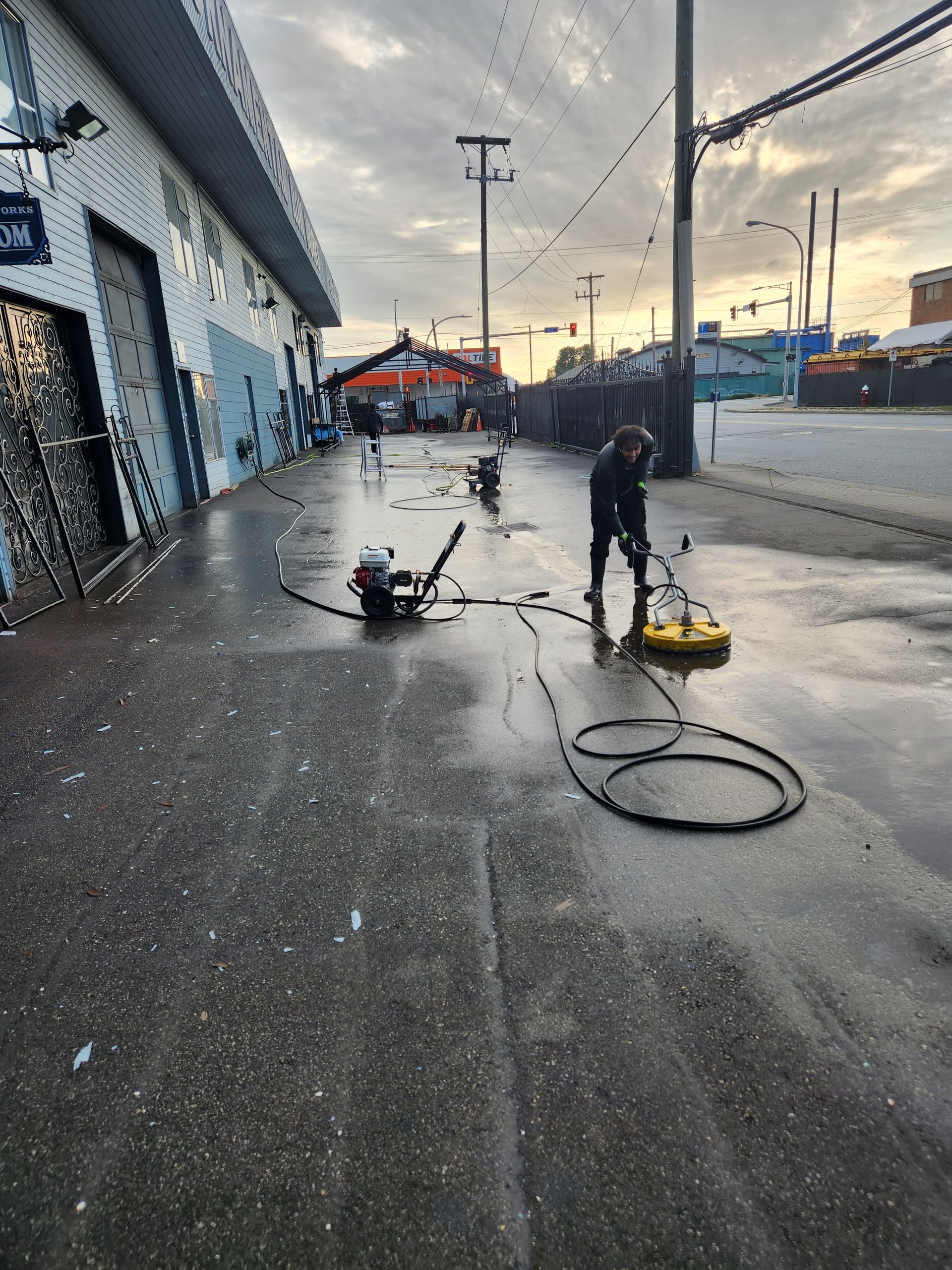 Person pressure washing a wet concrete sidewalk in front of a commercial building on a cloudy day.