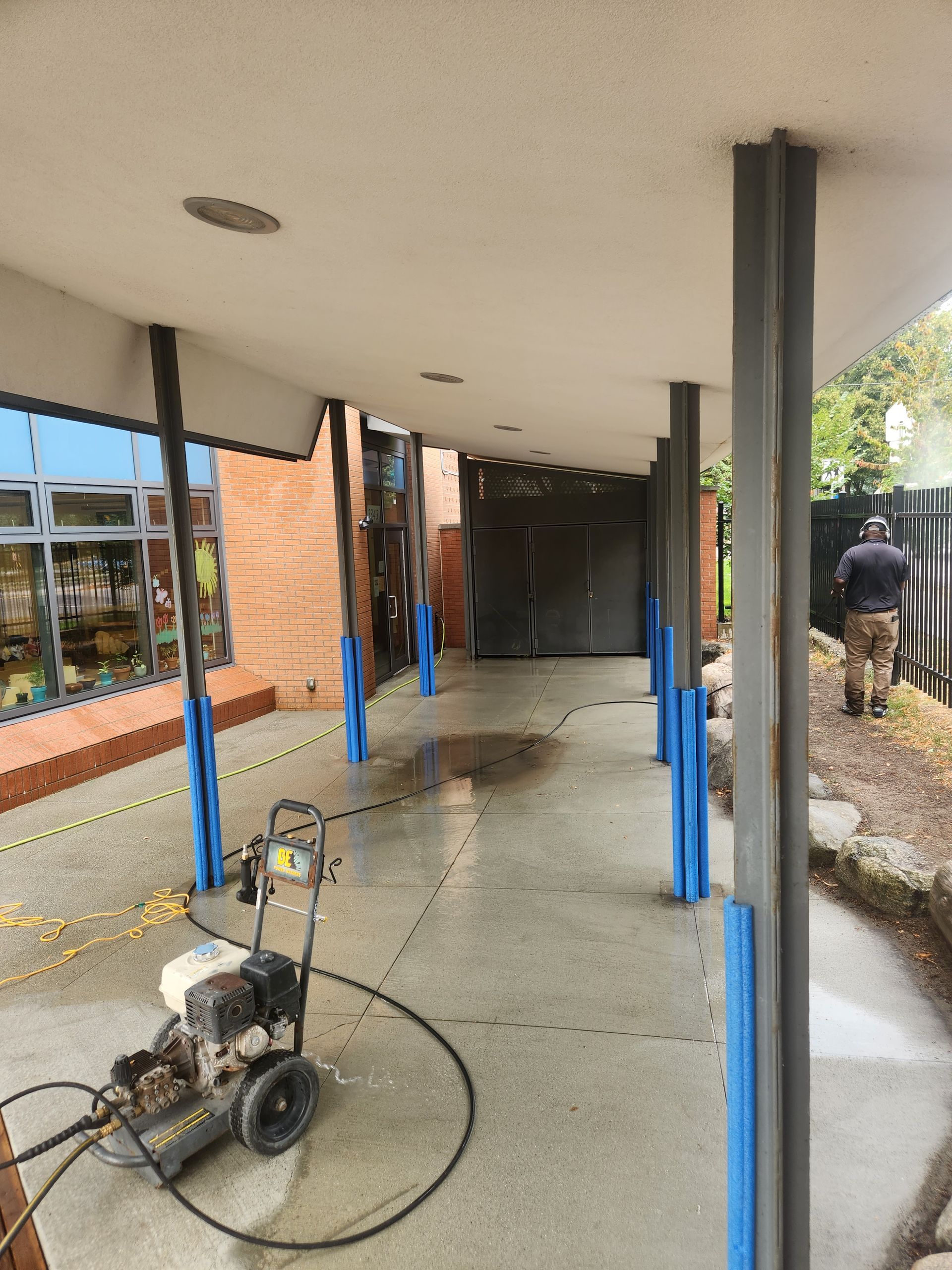 A person power washes a concrete walkway under a covered overhang, with blue-wrapped support pillars.