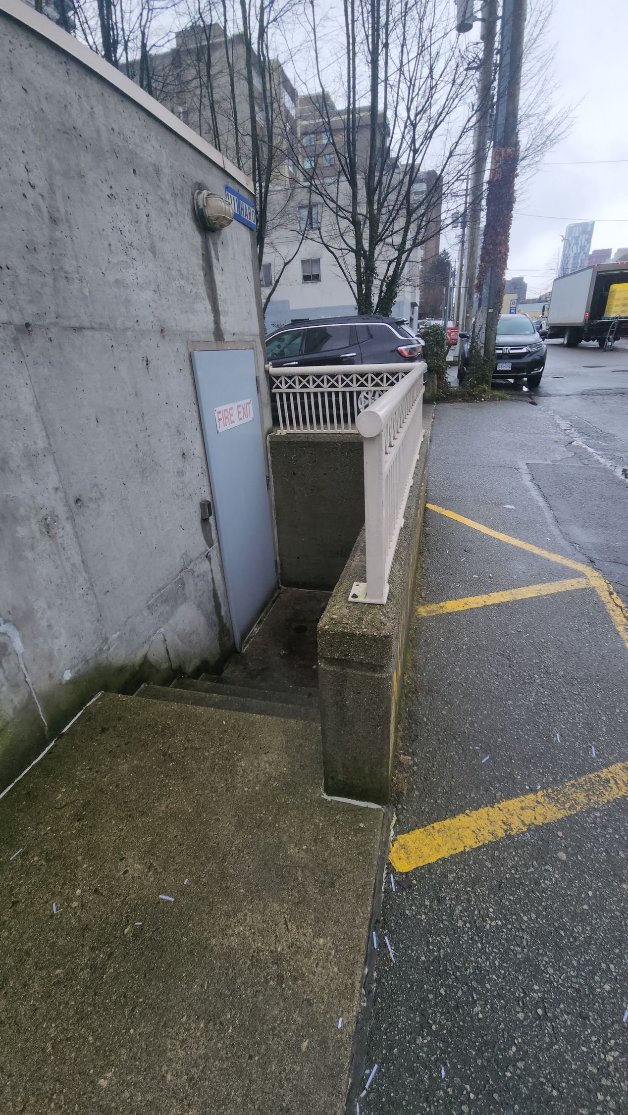 Concrete steps leading down to a doorway with a metal door, next to a curb and railing on a wet street.