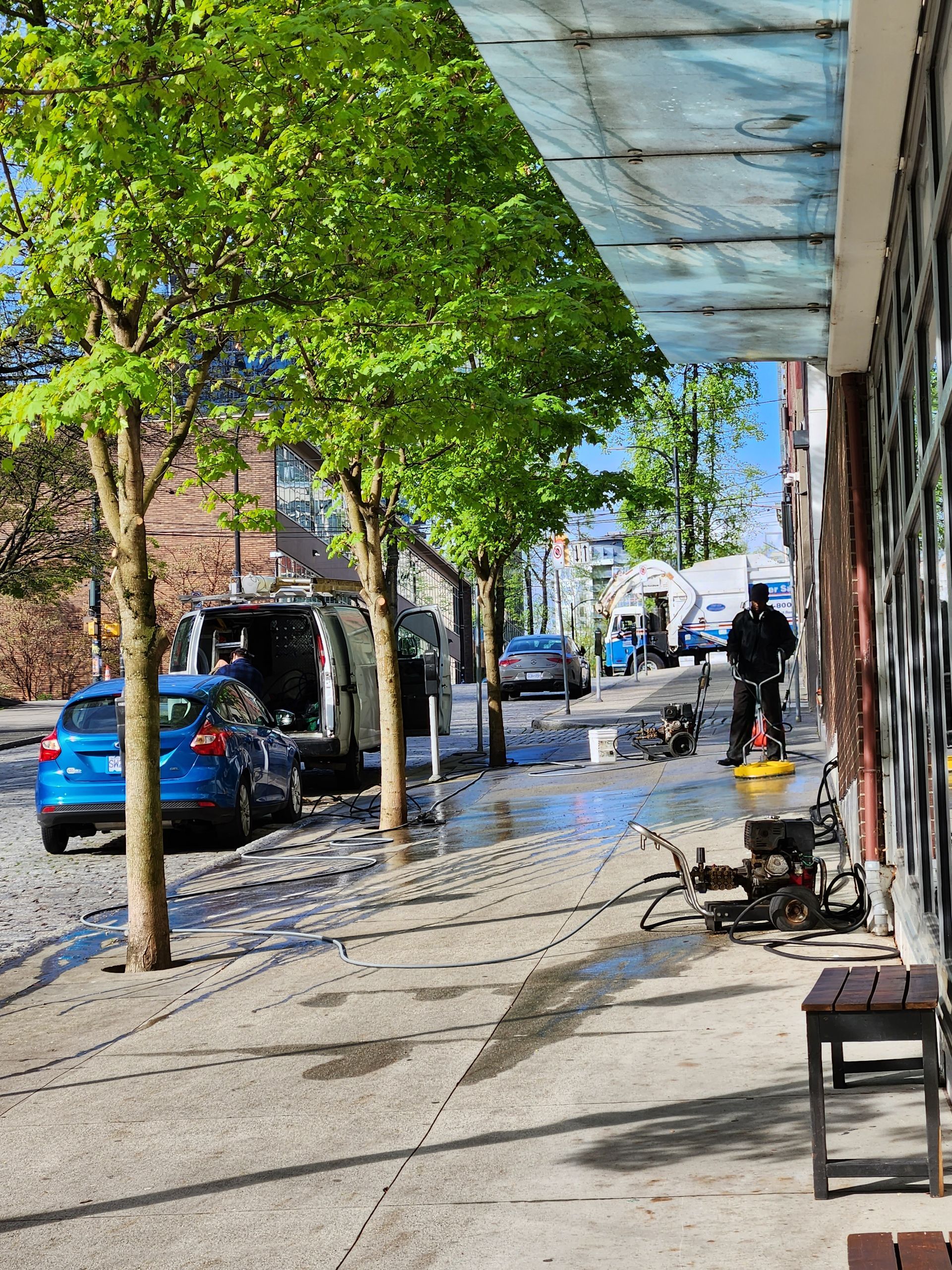 A person pressure washes a sidewalk next to trees and a parked blue car on a sunny day.