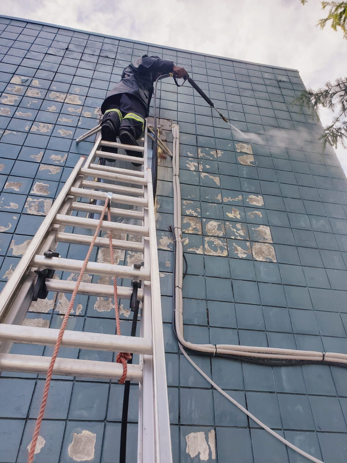 Person on a ladder power washing a blue tiled wall.
