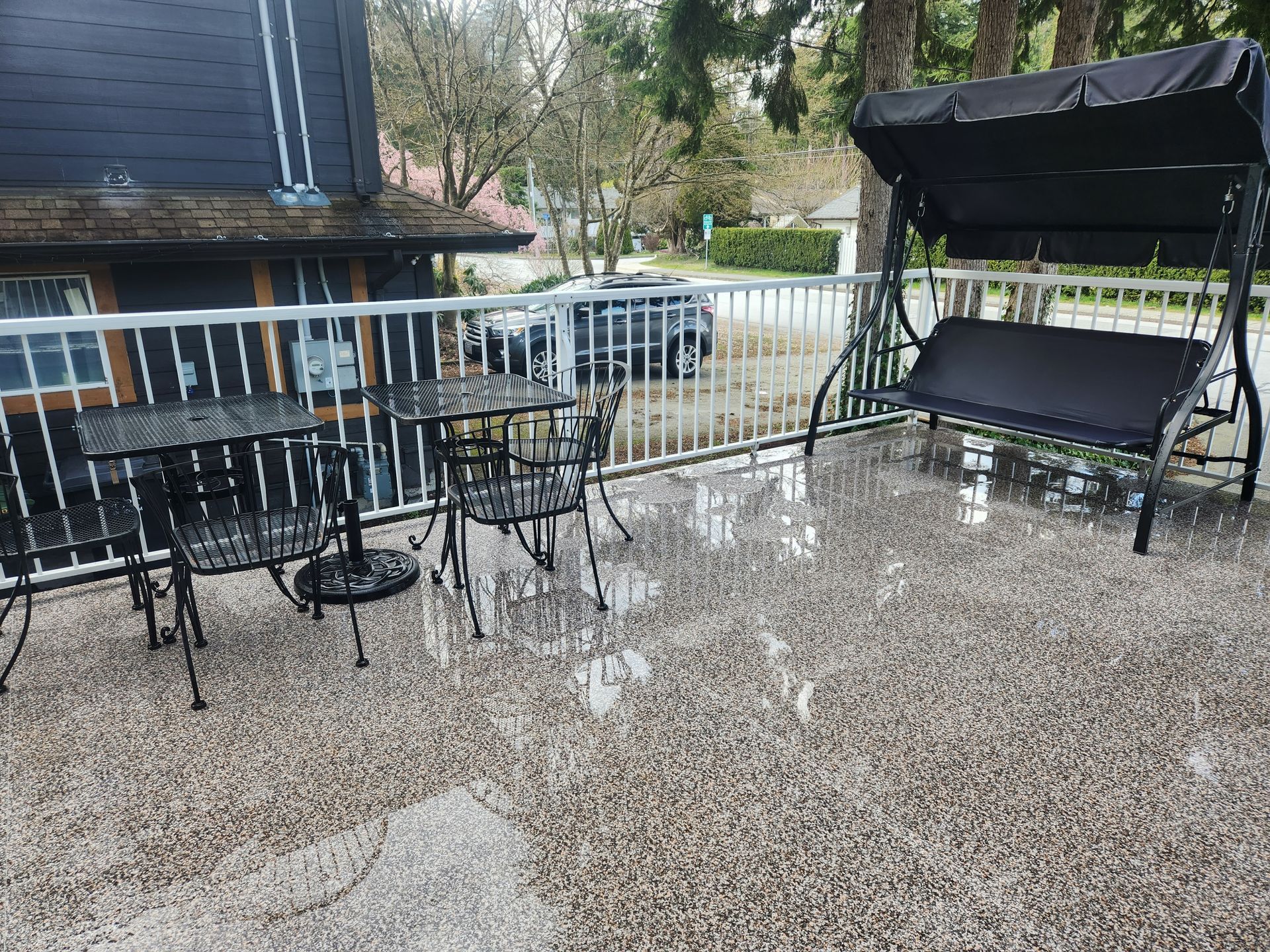 A rain-slicked patio with two small bistro tables, chairs, and a black porch swing set against a dark-sided building.