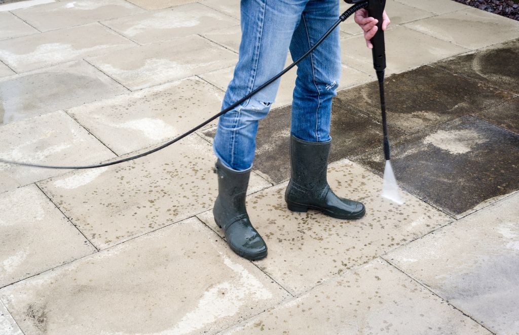 Person in jeans and boots pressure washing a stone patio.