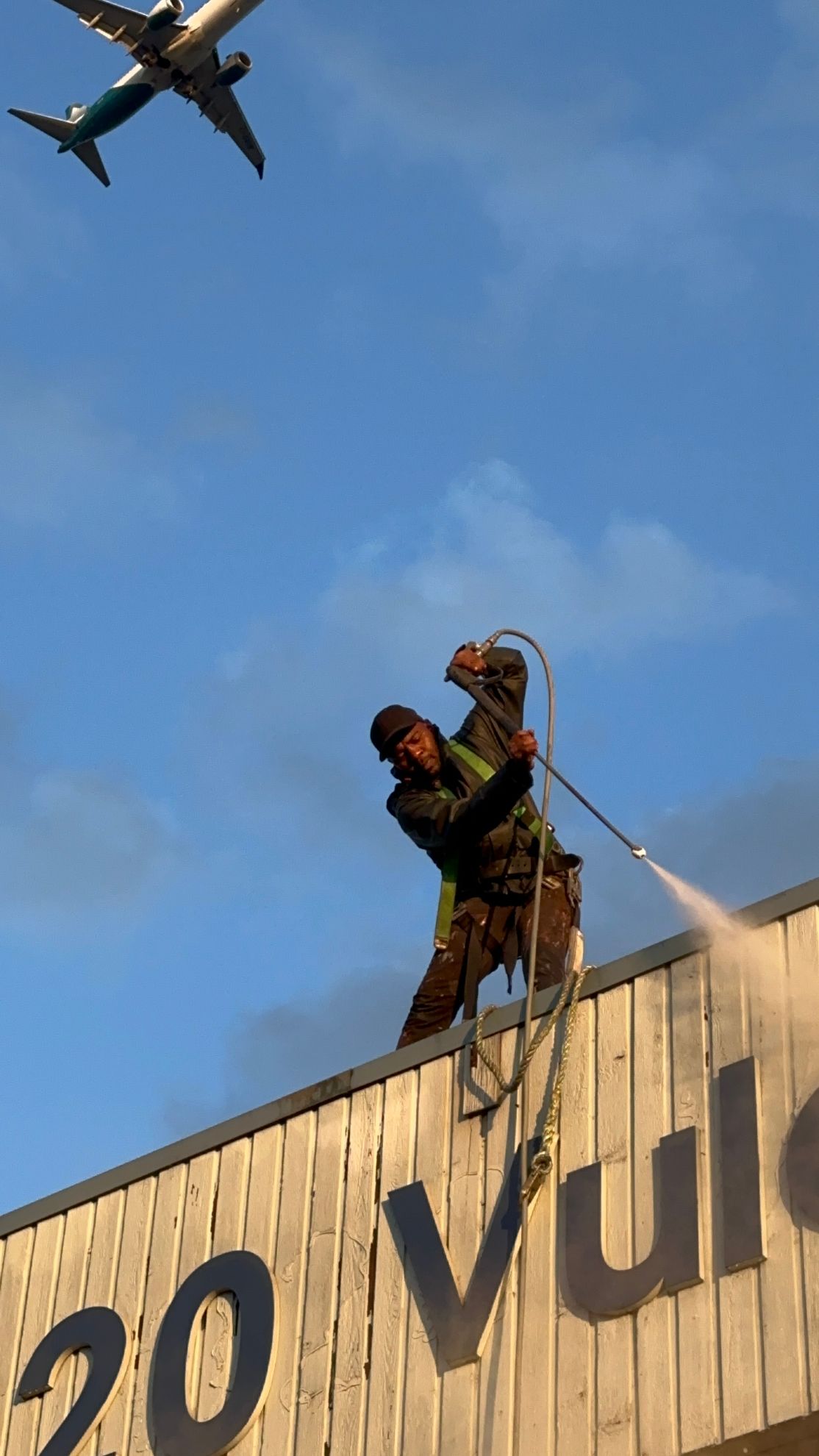 A person stands on top of a building, using a power washer to spray the wooden facade while an airplane flies overhead.