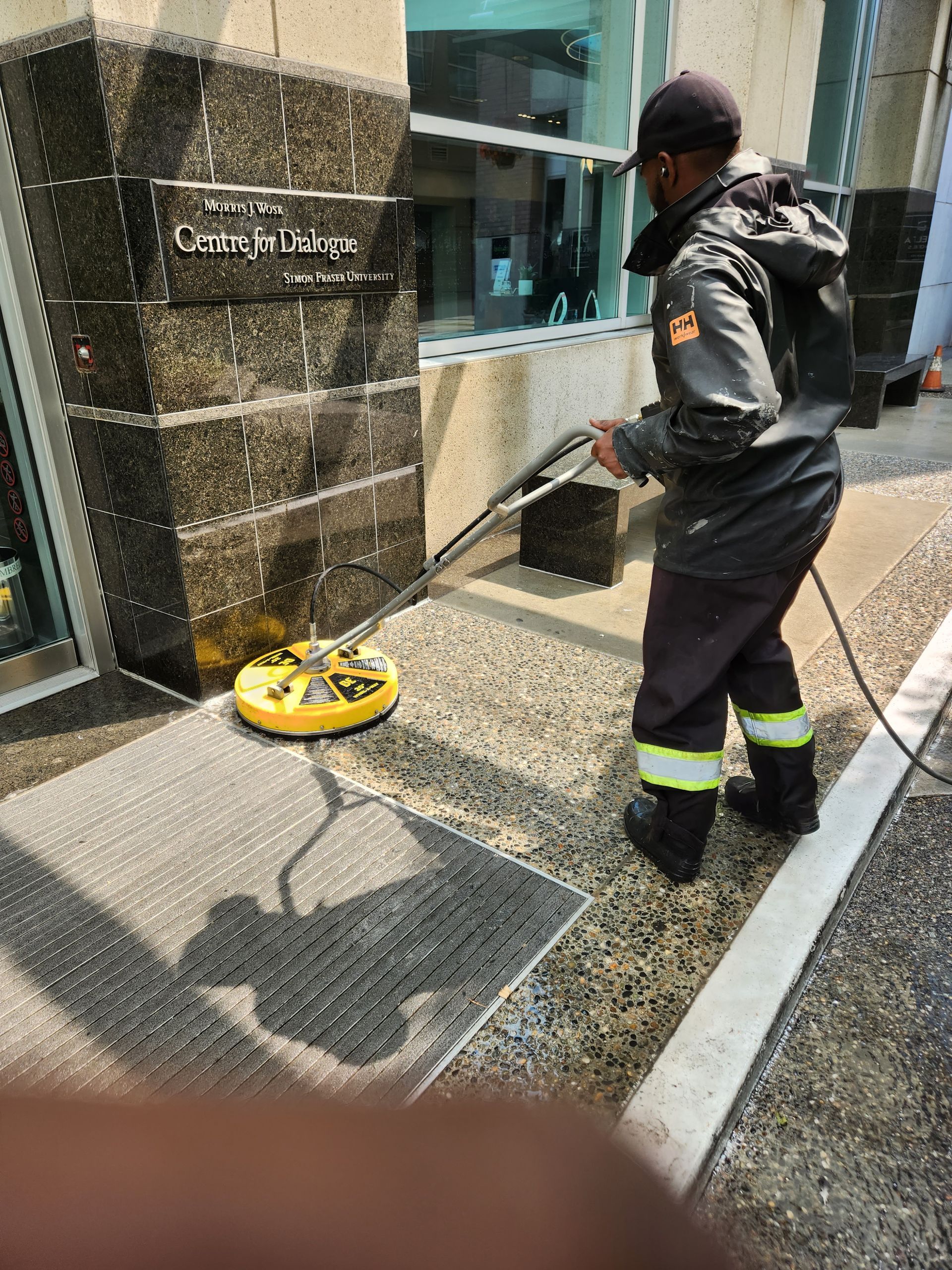 A worker in a dark jacket and pants uses a yellow surface cleaner to pressure wash a sidewalk outside a storefront.