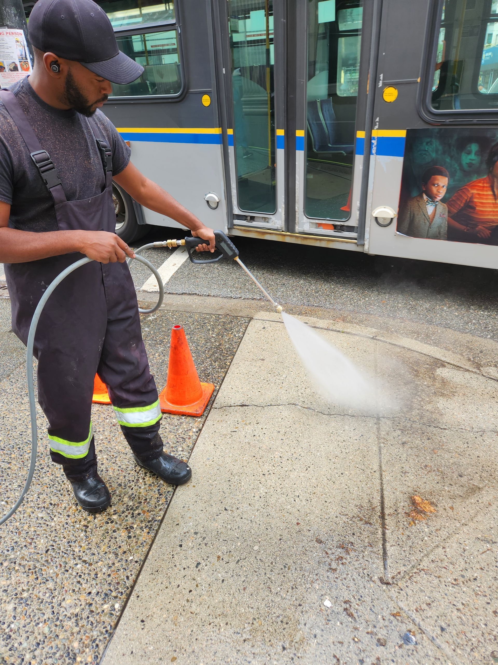 A person in work uniform uses a pressure washer to clean a concrete sidewalk next to a public transit bus.