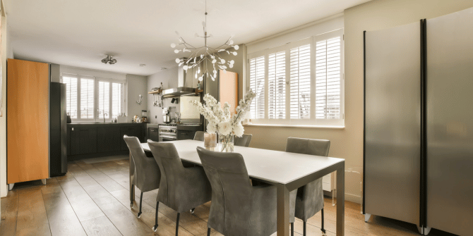 A dining room with a long table and chairs and a kitchen in the background.