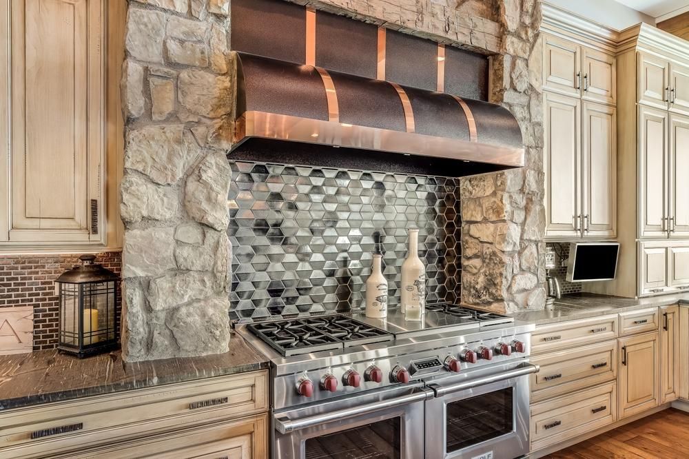 This kitchen has a rustic and natural feel, with stone finishes and warm wood accents. The cabinets are made of wood and have a distressed finish, which adds to the farmhouse-style aesthetic. The cabinet doors have a traditional panel design with visible knots and imperfections, contributing to the natural and rustic look of the kitchen. The countertops are made of natural stone, which adds a unique and organic element to the space. The backsplash is made of split octocagnol with a matte finish, which creates a subtle and neutral backdrop for the kitchen.