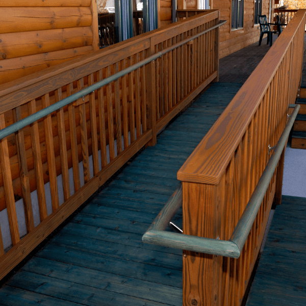 This image shows a residential property with a wheelchair ramp installed at the entrance. The ramp is long, gently sloping, and built with a sturdy metal structure. It starts from the driveway and leads up to the front door, providing a smooth transition for wheelchair users. The ramp has handrails on both sides for additional support and safety. There is also a small landing area at the top, allowing for easy maneuverability. The surface of the ramp appears to be textured, providing good traction to prevent slips and falls. The house itself is a single-story, suburban-style home with a brick facade and a large front yard. There's a driveway leading up to an attached garage. The yard is well-maintained, with a green lawn and a few small trees. Overall, the image illustrates an accessible home design that caters to individuals with mobility challenges.