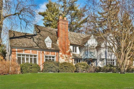 This image showcases a grand Tudor-style home located in Scarsdale, NY. The house is two-stories tall, featuring the distinctive half-timbered design and stucco walls that are typical of the Tudor architectural style. The roof is steep and multi-gabled, covered with shingles. The front facade of the house is symmetrical, with a centrally located arched front door. The door is made of dark wood and features ornate details, contributing to the home's old-world charm. It's flanked on both sides by bay windows with diamond-shaped leaded glass panes, enhancing the home's historical character. A brick pathway leads to the front entrance, and the lawn is beautifully manicured with a variety of mature trees and shrubs, adding to the property's curb appeal. The property appears to be situated in a quiet, residential neighborhood with similar homes in the background. Overall, the image depicts an elegant, well-preserved Tudor-style home, exuding charm and historical significance.