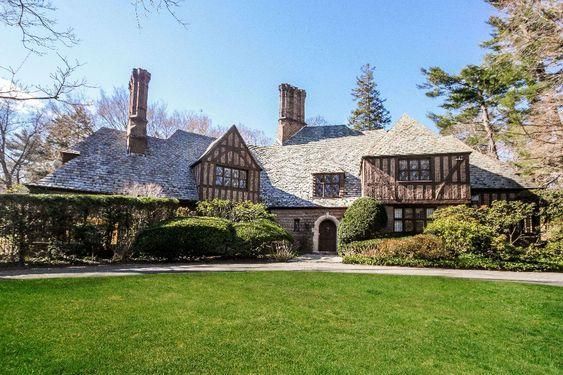 This image features a grand Tudor-style estate located in Scarsdale, NY. The house is an impressive multi-story structure, displaying the characteristic half-timbered design, brickwork, and steeply pitched gabled roofs that are typical of the Tudor architectural style. The facade of the house is symmetrically arranged, with a central front door and evenly spaced windows on each level. The windows are tall and narrow, featuring mullions that divide them into several panes, adding to the home's historical charm. The front door is arched and made of dark wood, offering a warm welcome. The estate is set on expansive, well-manicured grounds, complete with a large lawn, mature trees, and a wide driveway leading up to the house. The property appears to be located in a quiet, upscale residential neighborhood, with a clear blue sky overhead. Overall, the image presents an impressive, beautifully preserved Tudor-style estate, demonstrating a timeless architectural elegance and a high standard of living.