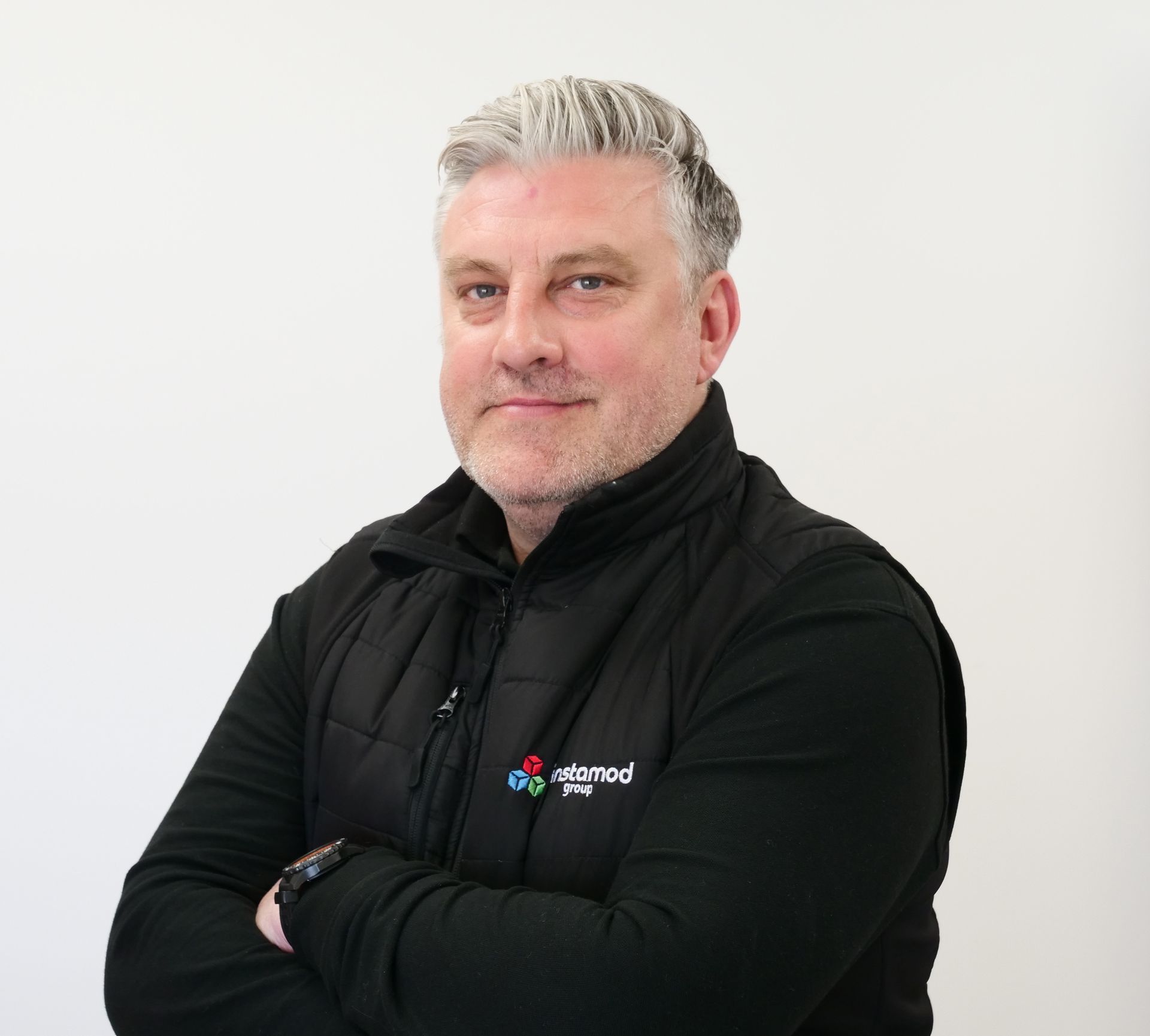 A man with a beard wearing a black vest is standing with arms crossed.